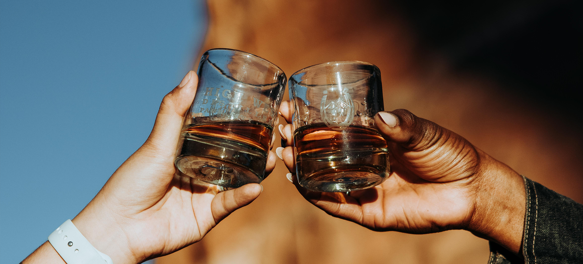 Two hands, holding High West highball glasses, clink together with a rugged cliffside behind them.