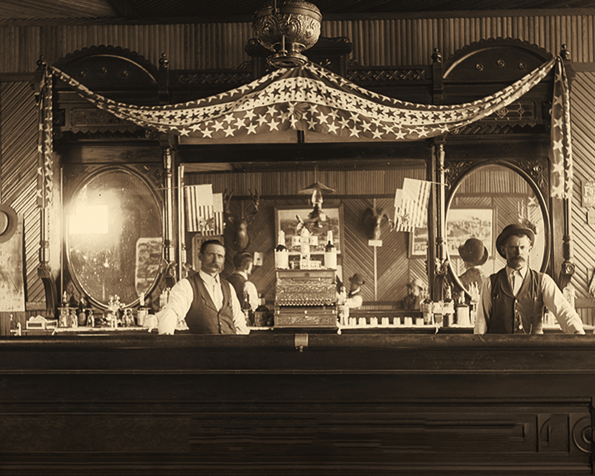 Two men stand behind the bar at a saloon from the 1800s. A festive banner drapes behind them.