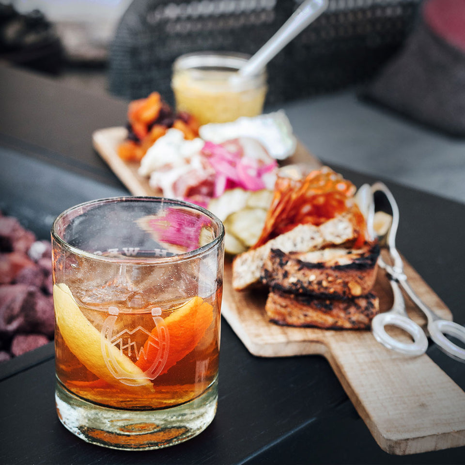 A glass of High West Double Rye with orange peel garnish on a table with BBQ snacks on a wooden cutting board.